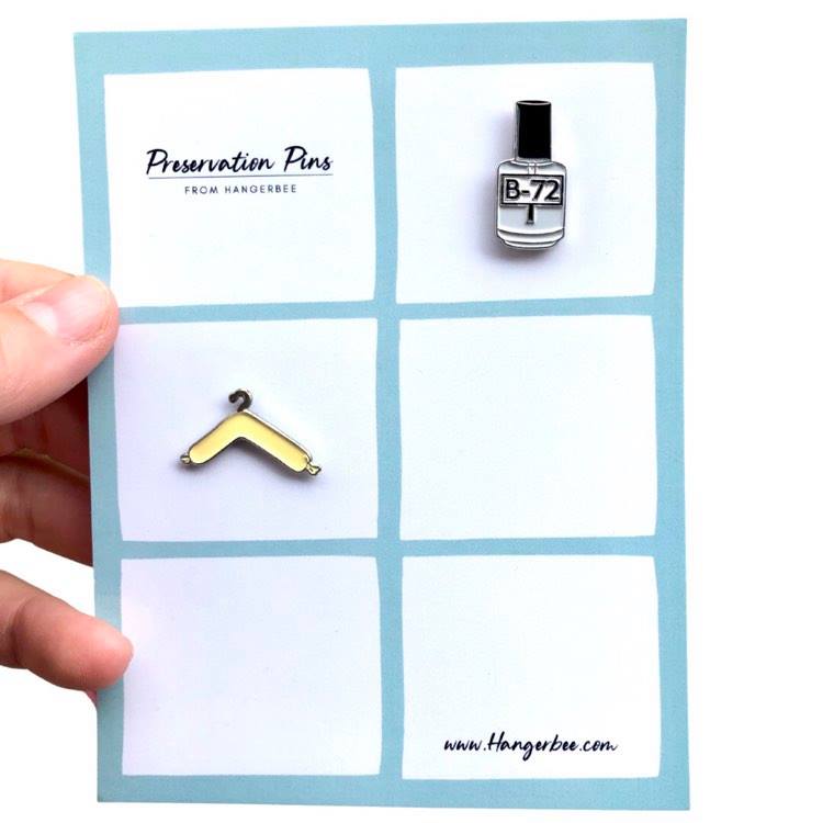 Photo of Hangerbee padded hanger's Collections Toolbox Lapel Pin set that includes a padded hanger pin, a b-72 bottle pin, a 1/4" photography scale pin, a hygrothermograph pin, and a pin of a hake brush and bone folder, all mounted on a card that's designed to look like an acid-free box lined with ethafoam. The card is on a small, cotton bag which is on a wood table.