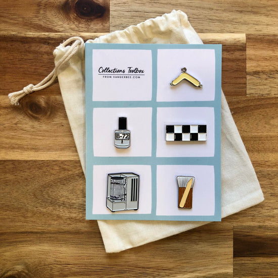 Photo of Hangerbee padded hanger's Collections Toolbox Lapel Pin set that includes a padded hanger pin, a b-72 bottle pin, a 1/4" photography scale pin, a hygrothermograph pin, and a pin of a hake brush and bone folder, all mounted on a card that's designed to look like an acid-free box lined with ethafoam. The card is on a small, cotton bag which is on a wood table.