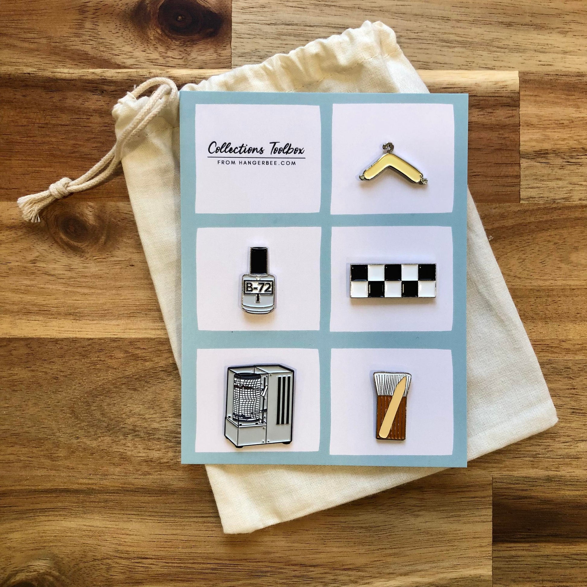 Photo of Hangerbee padded hanger's Collections Toolbox Lapel Pin set that includes a padded hanger pin, a b-72 bottle pin, a 1/4" photography scale pin, a hygrothermograph pin, and a pin of a hake brush and bone folder, all mounted on a card that's designed to look like an acid-free box lined with ethafoam. The card is on a small, cotton bag which is on a wood table.