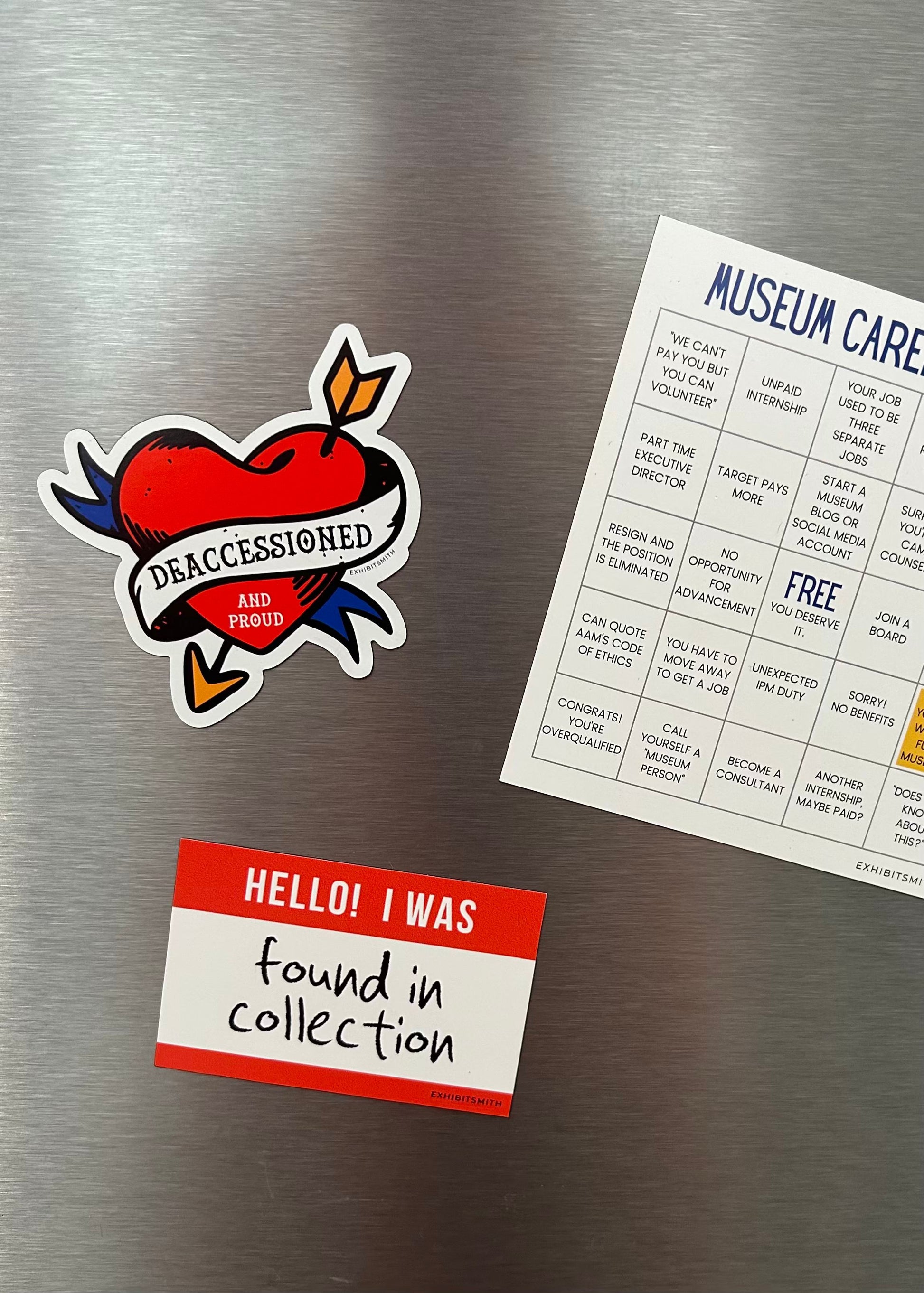 Hangerbee padded clothing hangers has developed magnets, stickers, keychains, pins, and more, with museum collections professionals in mind. This image shows a Deaccessioned and Proud magnet and a Found in Collection magnet holding up a square Hangerbee business card on a refrigerator.