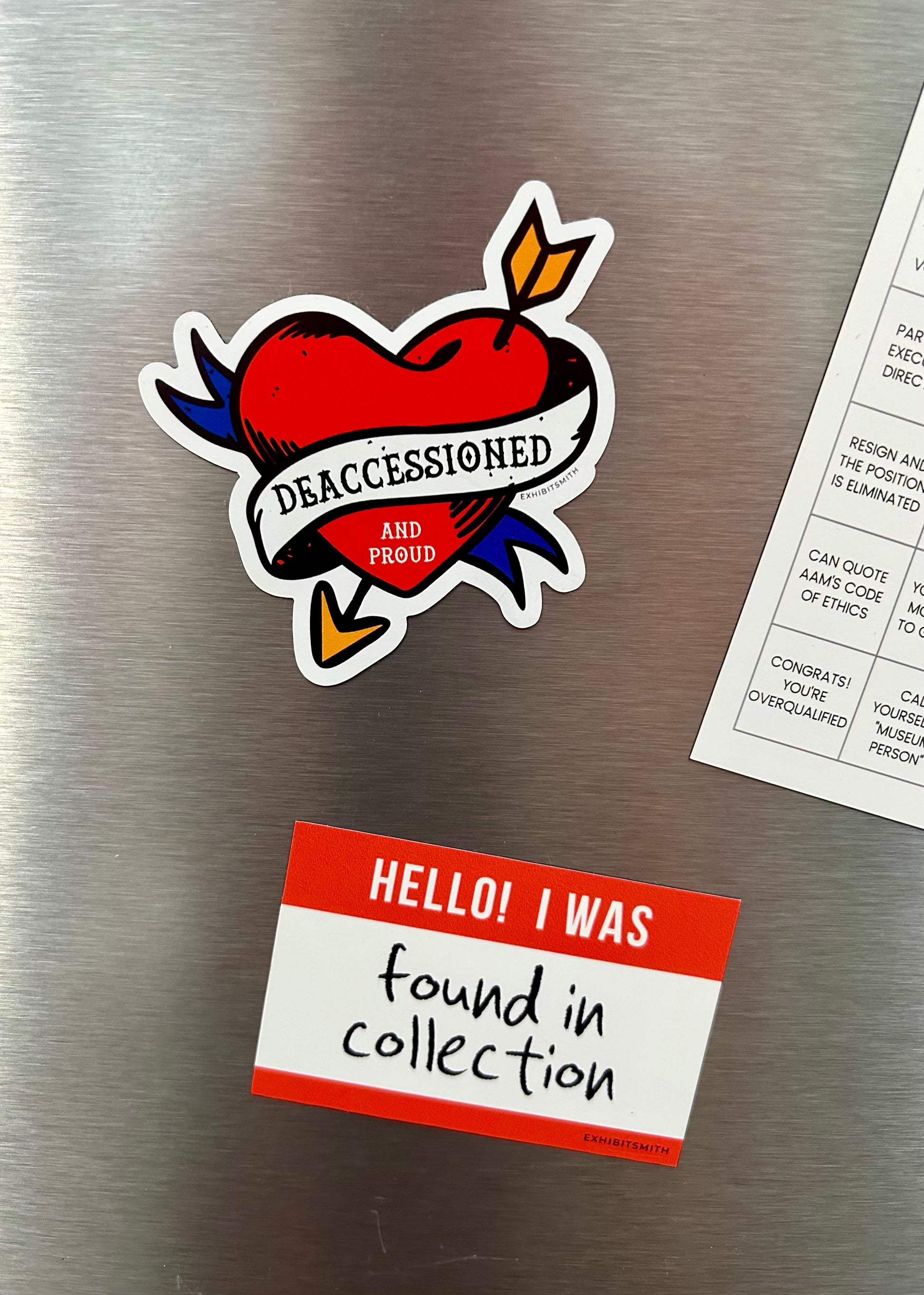 Hangerbee padded clothing hangers has developed magnets, stickers, keychains, pins, and more, with museum collections professionals in mind. This image shows a Deaccessioned and Proud magnet and a Found in Collection magnet holding up a square Hangerbee business card on a refrigerator.