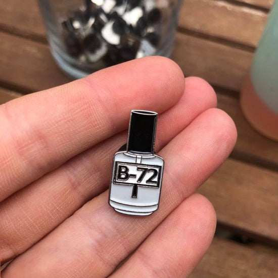 Photo of Hangerbee padded hanger's Collections Toolbox Lapel Pin of a Paraloid / Acryloid B-72 bottle on a card that's designed to look like an acid-free box lined with ethafoam. The card is on a small, cotton bag which is on a wood table.