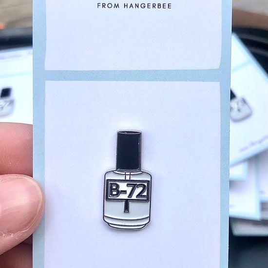 Photo of Hangerbee padded hanger's Collections Toolbox Lapel Pin of a Paraloid / Acryloid B-72 bottle on a card that's designed to look like an acid-free box lined with ethafoam. The card is on a small, cotton bag which is on a wood table.
