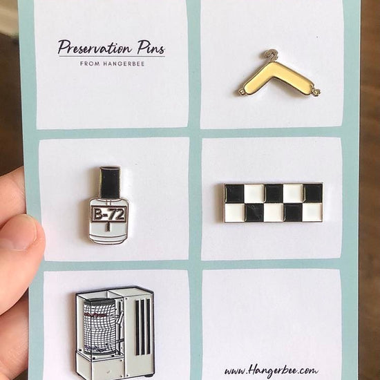 Photo of Hangerbee padded hanger's Collections Toolbox Lapel Pin set that includes a padded hanger pin, a b-72 bottle pin, a 1/4" photography scale pin, a hygrothermograph pin, and a pin of a hake brush and bone folder, all mounted on a card that's designed to look like an acid-free box lined with ethafoam. The card is on a small, cotton bag which is on a wood table.