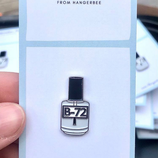 Photo of Hangerbee padded hanger's Collections Toolbox Lapel Pin of a paraloid / acryloid b-72 bottle on a card that's designed to look like an acid-free box lined with ethafoam. The card is on a small, cotton bag which is on a wood table.