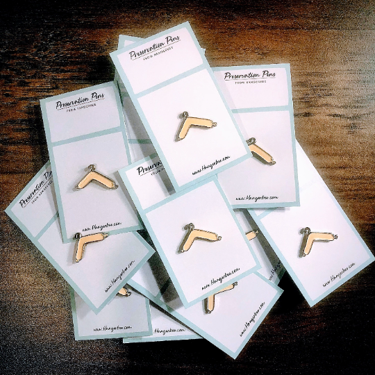 Photo of Hangerbee padded hanger's Collections Toolbox Lapel Pin of padded hanger pins on cards that are designed to look like an acid-free box lined with ethafoam. The card is on a small, cotton bag which is on a wood table.