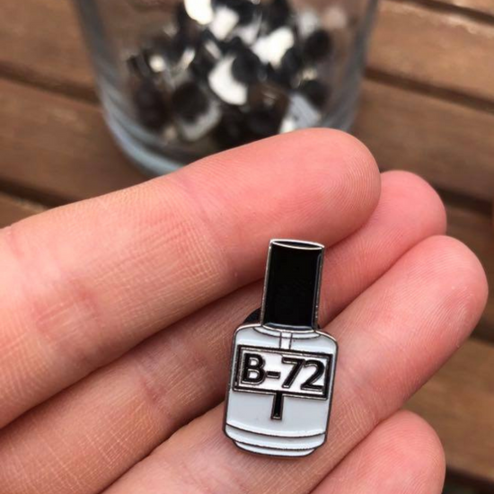 Photo of Hangerbee padded hanger's Collections Toolbox Lapel Pin of a paraloid / acryloid B-72 bottle on a card that's designed to look like an acid-free box lined with ethafoam. The card is on a small, cotton bag which is on a wood table.