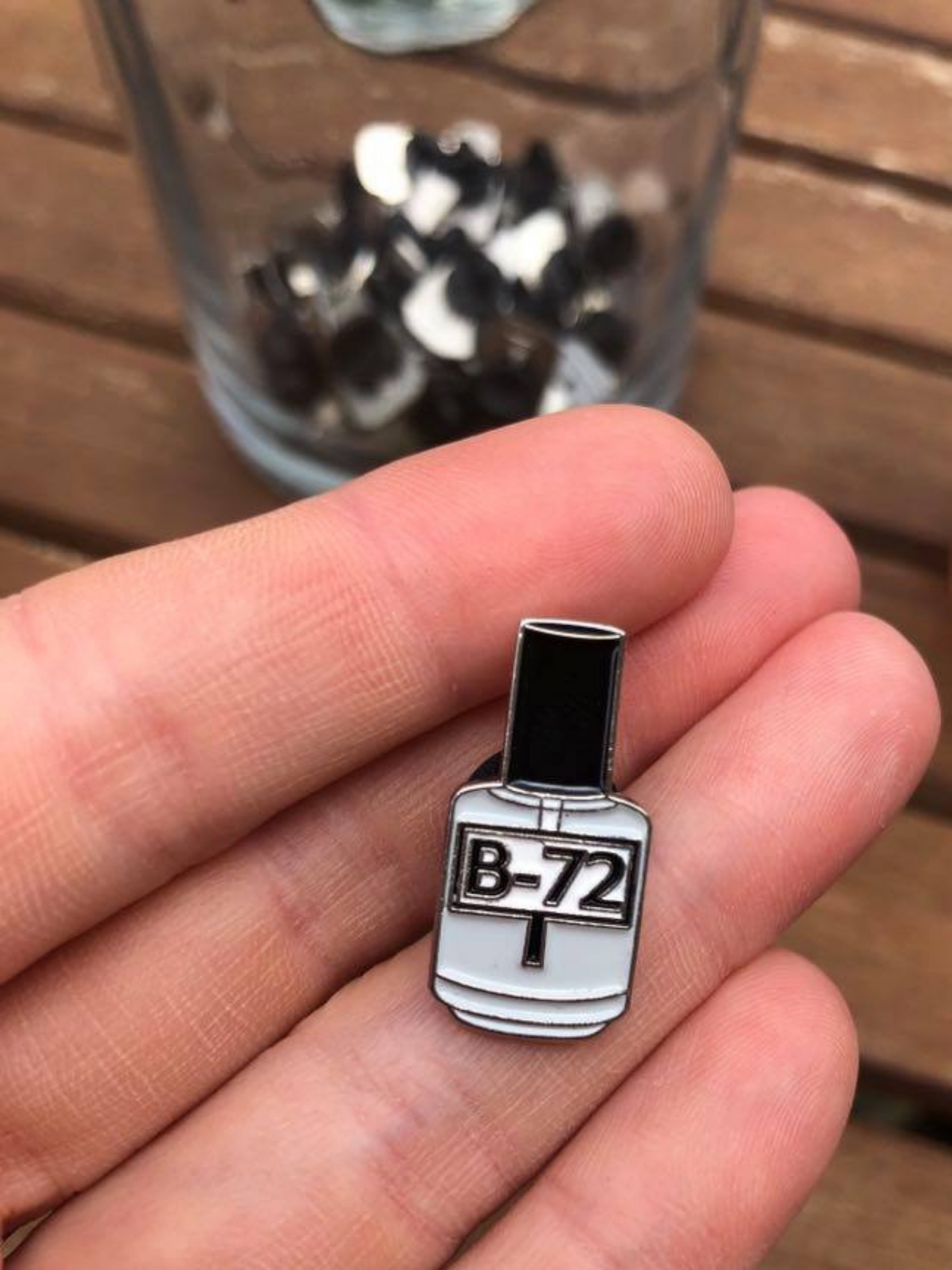 Photo of Hangerbee padded hanger's Collections Toolbox Lapel Pin of a paraloid / acryloid B-72 bottle on a card that's designed to look like an acid-free box lined with ethafoam. The card is on a small, cotton bag which is on a wood table.