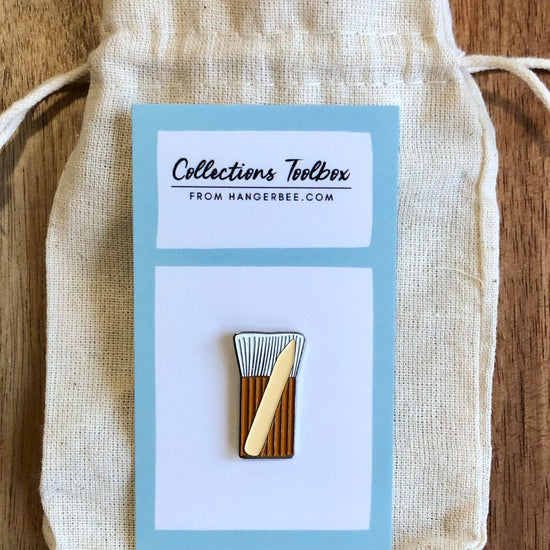 Photo of Hangerbee padded hanger's Collections Toolbox Lapel Pin of a hake brush and bone folder on  card that's designed to look like an acid-free box lined with ethafoam. The card is on a small, cotton bag which is on a wood table.