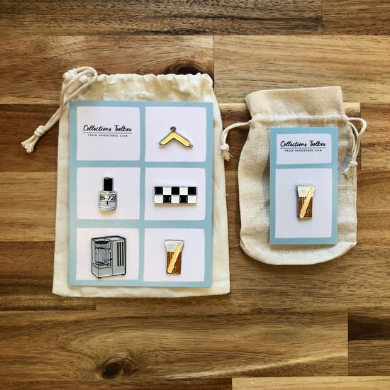 Photo of Hangerbee padded hanger's Collections Toolbox Lapel Pin set that includes a padded hanger pin, a b-72 bottle pin, a 1/4" photography scale pin, a hygrothermograph pin, and a pin of a hake brush and bone folder, all mounted on a card that's designed to look like an acid-free box lined with ethafoam. The card is on a small, cotton bag which is on a wood table.