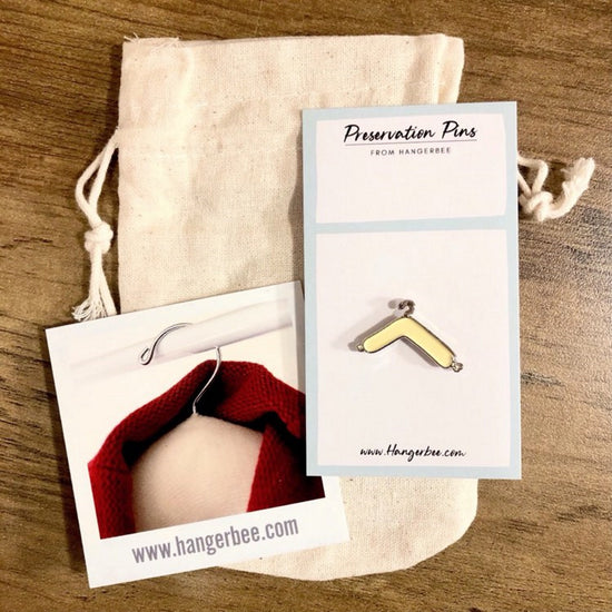 Photo of Hangerbee padded hanger's Collections Toolbox Lapel Pin of a hangerbee padded hanger on a card that's designed to look like an acid-free box lined with ethafoam. The card is on a small, cotton bag which is on a wood table.