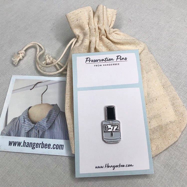 Photo of Hangerbee padded hanger's Collections Toolbox Lapel Pin of a Paraloid / acryloid B72 on a card that's designed to look like an acid-free box lined with ethafoam. The card is on a small, cotton bag which is on a wood table.