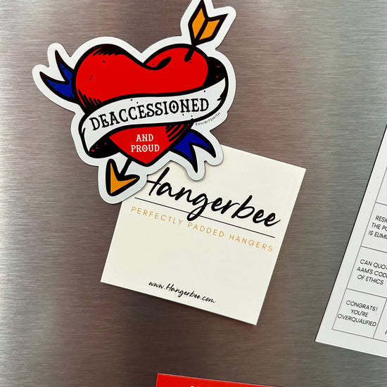 Hangerbee padded clothing hangers has developed magnets, stickers, keychains, pins, and more, with museum collections professionals in mind. This image shows a Deaccessioned and Proud magnet and a Found in Collection magnet holding up a square Hangerbee business card on a refrigerator. 