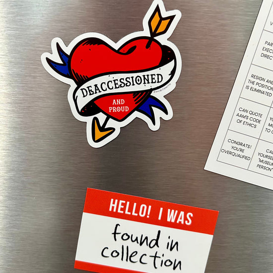 Hangerbee padded clothing hangers has developed magnets, stickers, keychains, pins, and more, with museum collections professionals in mind. This image shows a Deaccessioned and Proud magnet and a Found in Collection magnet holding up a square Hangerbee business card on a refrigerator. 