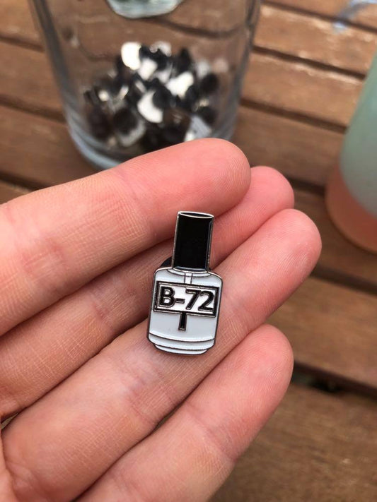 Photo of Hangerbee padded hanger's Collections Toolbox Lapel Pin of a Paraloid / Acryloid B-72 bottle on a card that's designed to look like an acid-free box lined with ethafoam. The card is on a small, cotton bag which is on a wood table.