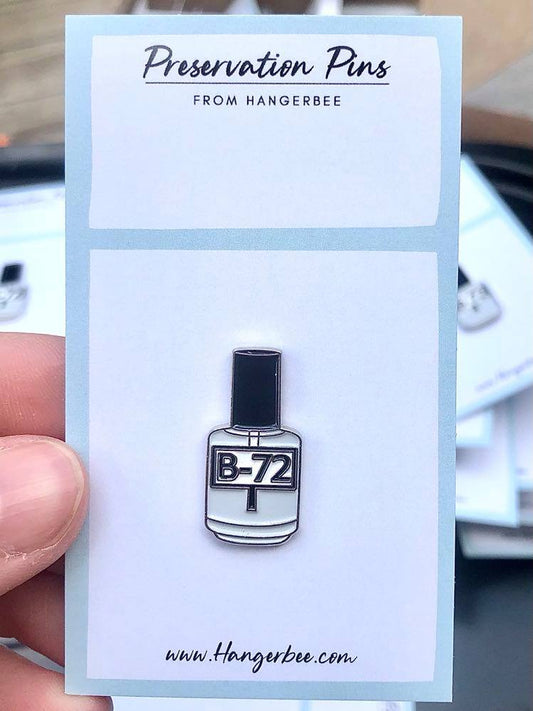 Photo of Hangerbee padded hanger's Collections Toolbox Lapel Pin of a Paraloid / Acryloid B-72 bottle on a card that's designed to look like an acid-free box lined with ethafoam. The card is on a small, cotton bag which is on a wood table.