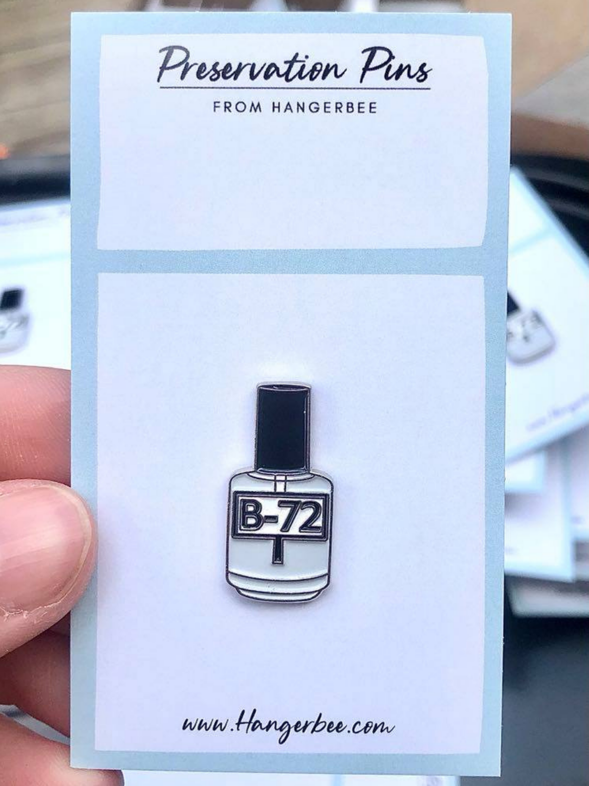 Photo of Hangerbee padded hanger's Collections Toolbox Lapel Pin of a paraloid / acryloid b-72 bottle on a card that's designed to look like an acid-free box lined with ethafoam. The card is on a small, cotton bag which is on a wood table.
