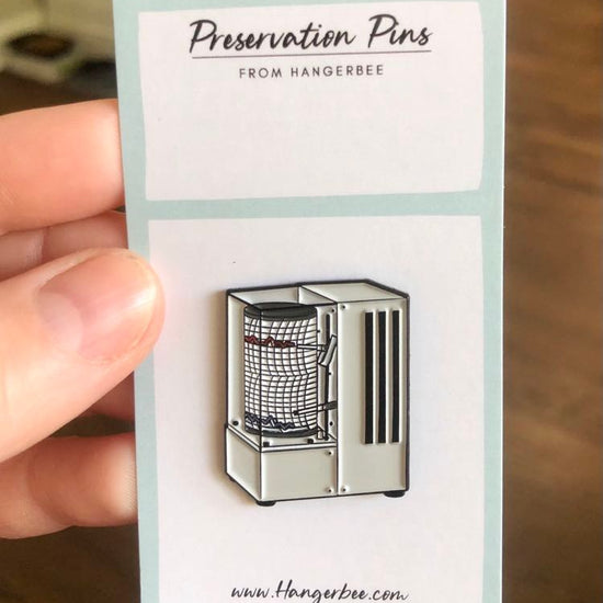 Photo of Hangerbee padded hanger's Collections Toolbox Lapel Pin of a hygrothermograph on a card that's designed to look like an acid-free box lined with ethafoam. The card is on a small, cotton bag which is on a wood table.