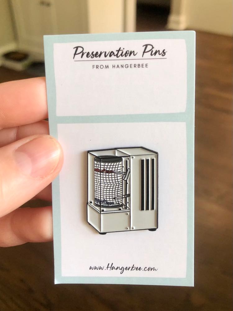 Photo of Hangerbee padded hanger's Collections Toolbox Lapel Pin of a hygrothermograph on a card that's designed to look like an acid-free box lined with ethafoam. The card is on a small, cotton bag which is on a wood table.