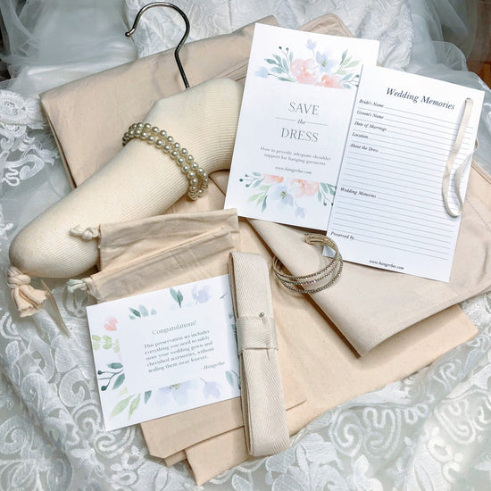 image featuring items in hangerbee padded hanger's save the dress, wedding dress preservation kit. The kit includes an heirloom safe, preservation quality padded clothes hanger, a large cotton garment bag, a large, zippered, cotton accessory bag and two small cotton drawstring bags, yards of cotton twill take, a wedding memories memory card, and instructions.