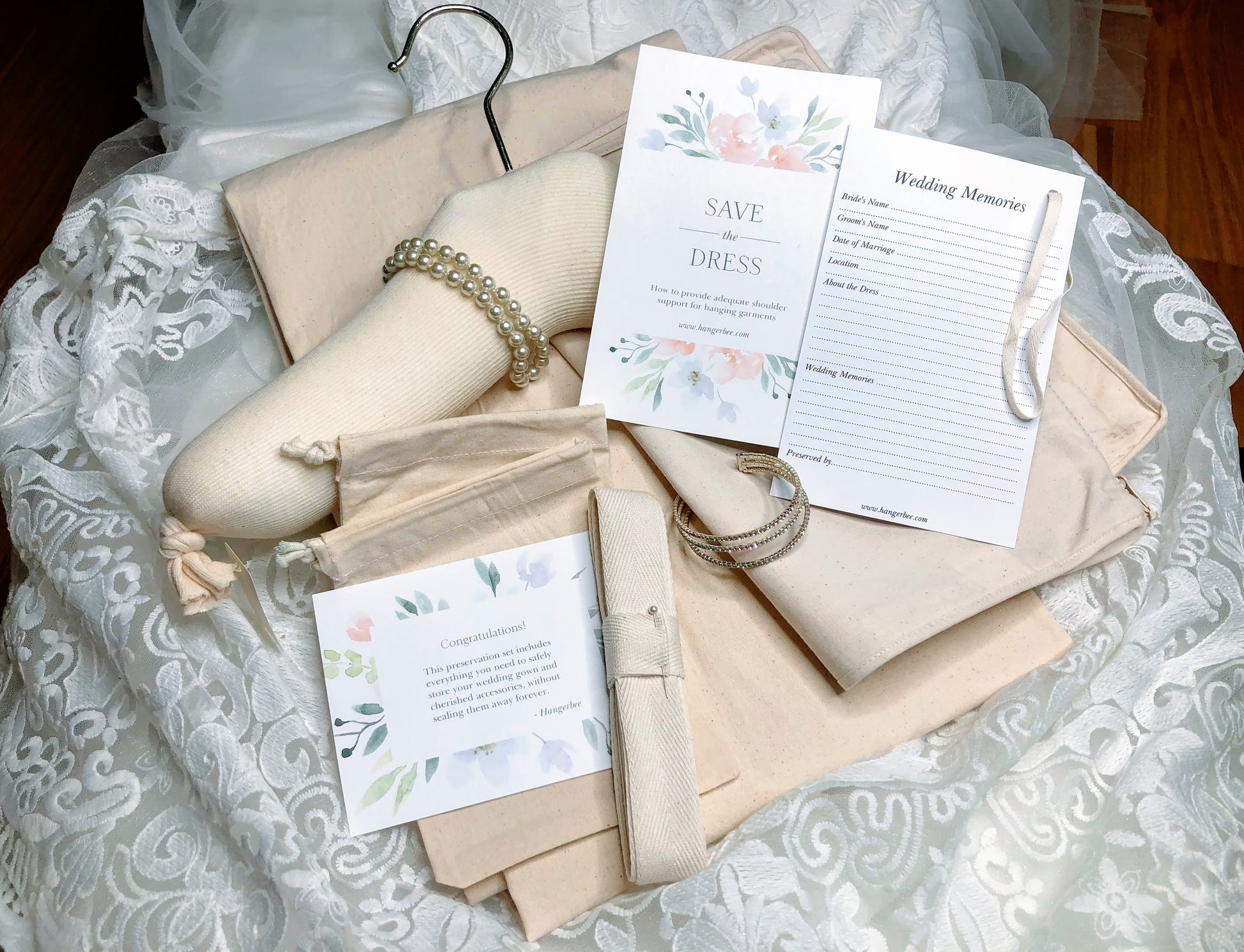 image featuring items in hangerbee padded hanger's save the dress, wedding dress preservation kit. The kit includes an heirloom safe, preservation quality padded clothes hanger, a large cotton garment bag, a large, zippered, cotton accessory bag and two small cotton drawstring bags, yards of cotton twill take, a wedding memories memory card, and instructions.