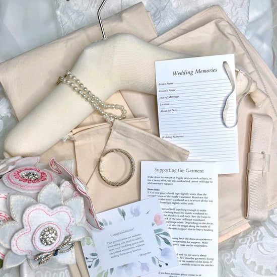image featuring items in hangerbee padded hanger's save the dress, wedding dress preservation kit. The kit includes an heirloom safe, preservation quality padded clothes hanger, a large cotton garment bag, a large, zippered, cotton accessory bag and two small cotton drawstring bags, yards of cotton twill take, a wedding memories memory card, and instructions.