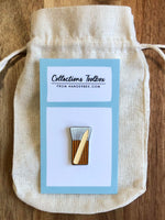 Photo of Hangerbee padded hanger's Collections Toolbox Lapel Pin of a hake brush and bone folder on  card that's designed to look like an acid-free box lined with ethafoam. The card is on a small, cotton bag which is on a wood table.