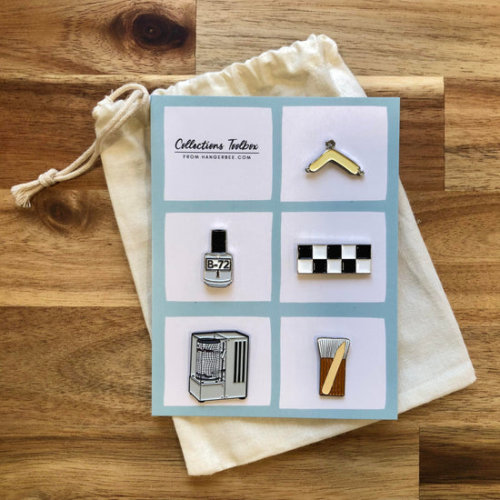 Photo of Hangerbee padded hanger's Collections Toolbox Lapel Pin set that includes a padded hanger pin, a b-72 bottle pin, a 1/4" photography scale pin, a hygrothermograph pin, and a pin of a hake brush and bone folder, all mounted on a card that's designed to look like an acid-free box lined with ethafoam. The card is on a small, cotton bag which is on a wood table.