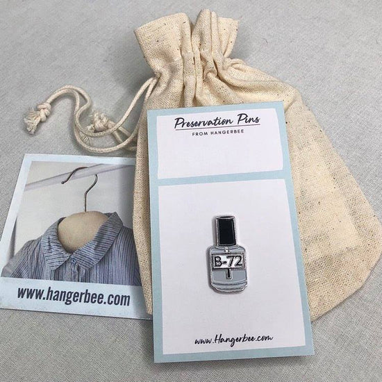 Photo of Hangerbee padded hanger's Collections Toolbox Lapel Pin of a Paraloid / acryloid B72 on a card that's designed to look like an acid-free box lined with ethafoam. The card is on a small, cotton bag which is on a wood table.