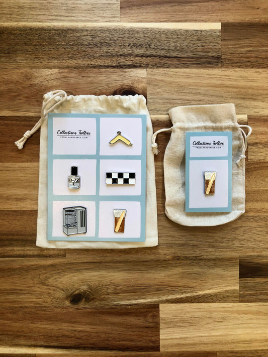 Photo of Hangerbee padded hanger's Collections Toolbox Lapel Pin set that includes a padded hanger pin, a b-72 bottle pin, a 1/4" photography scale pin, a hygrothermograph pin, and a pin of a hake brush and bone folder, all mounted on a card that's designed to look like an acid-free box lined with ethafoam. The card is on a small, cotton bag which is on a wood table.